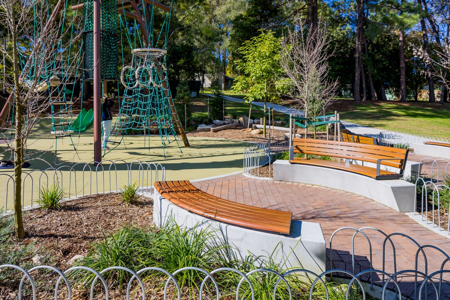 Curved timber bench on a brick-paved seating area beside a playground with rope climbing net, native plants and a low looped fence.