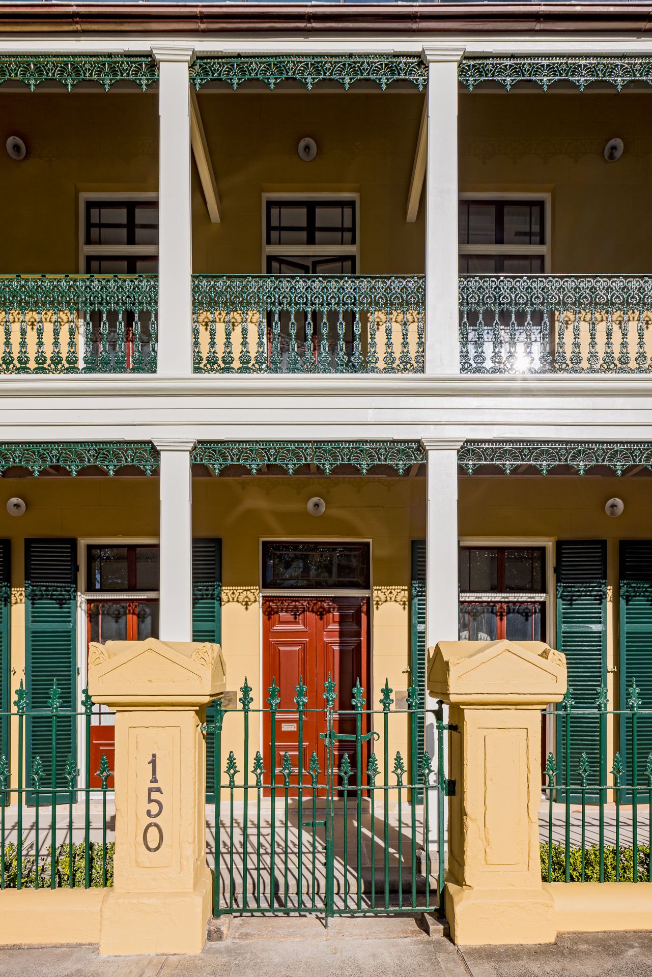 Historic two-storey terrace house with lacework iron balconies, red front door, green shutters and number 150 on the gate pillar.