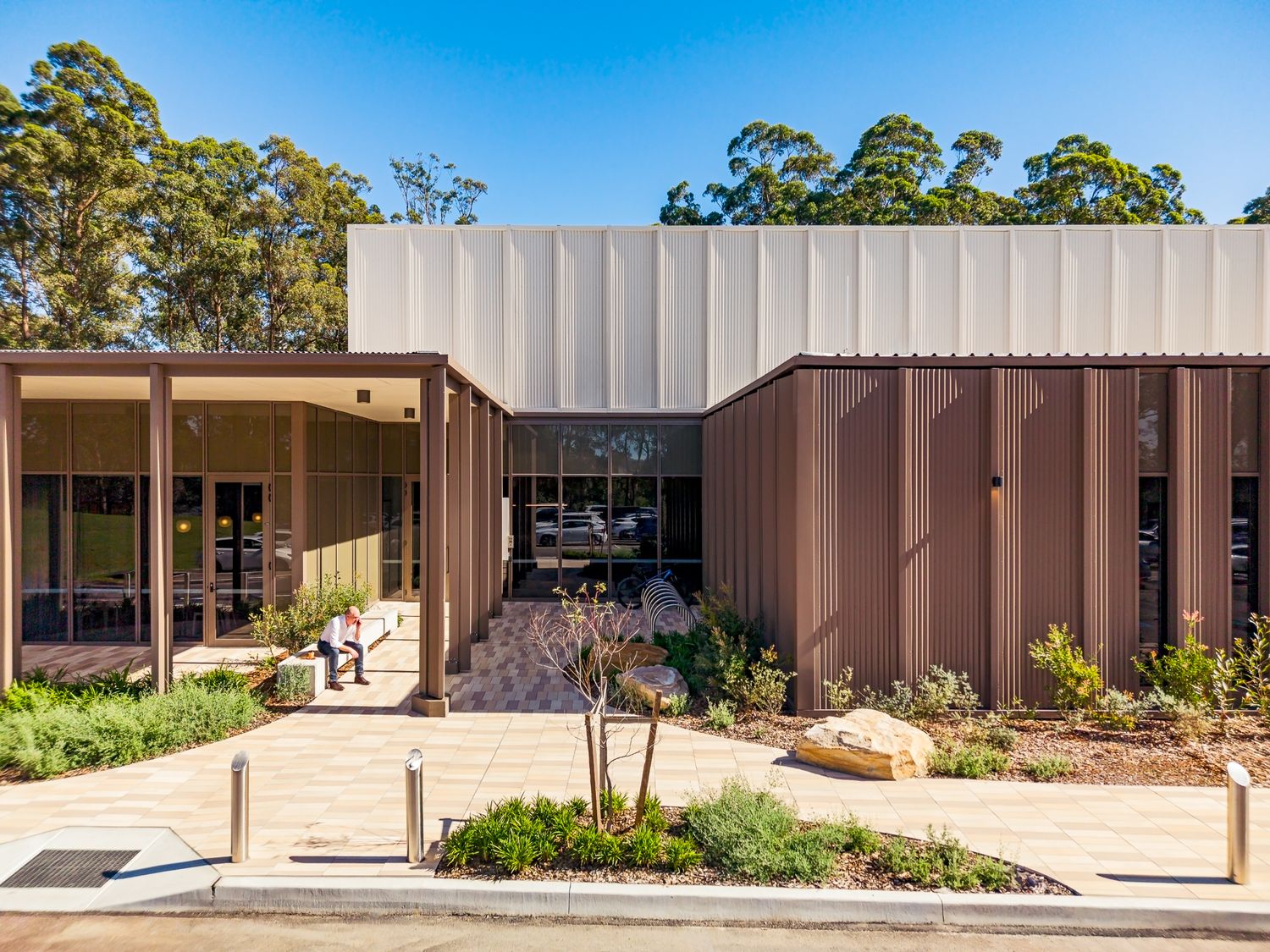 Entrance of a modern building with brown metal cladding, glass doors and a paved path lined with native plants under tall gum trees.