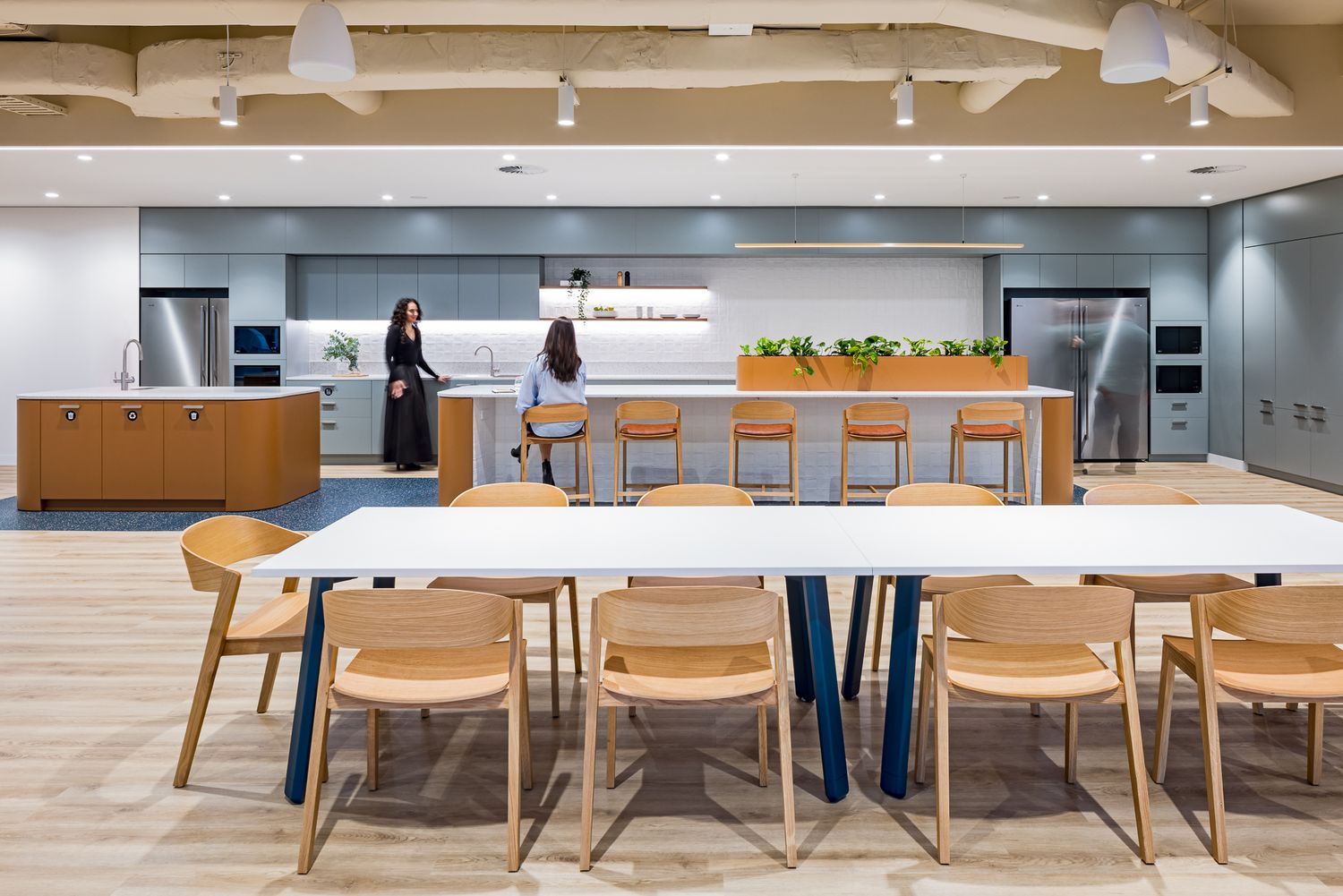 Two employees stand near a kitchen island in a modern office pantry with a white communal table and wooden dining chairs.