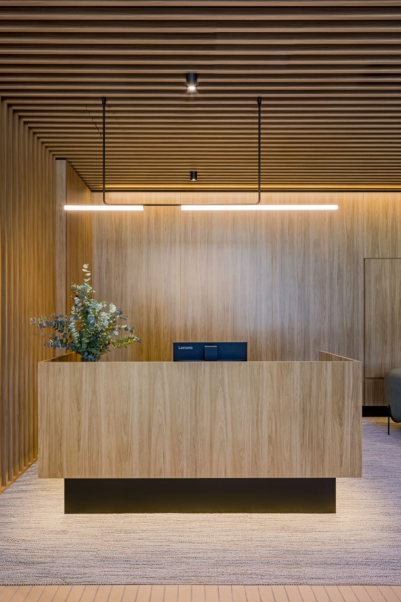 Timber reception desk with a eucalyptus plant, computer monitor and linear pendant lighting against a warm wood-slat feature wall.