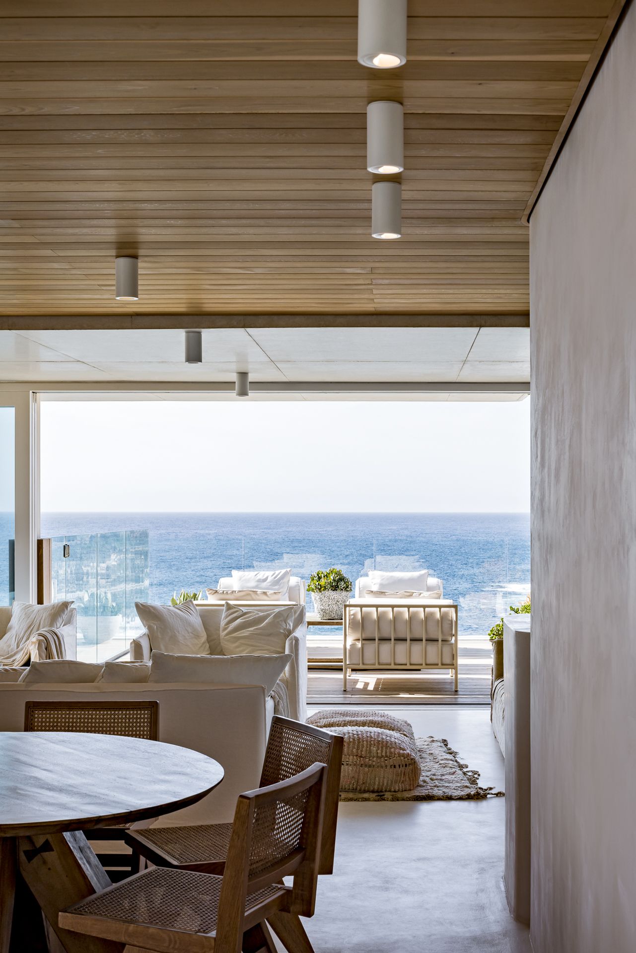 Dining area with rattan chairs and a timber ceiling opening onto a terrace with outdoor seating and a wide ocean horizon.