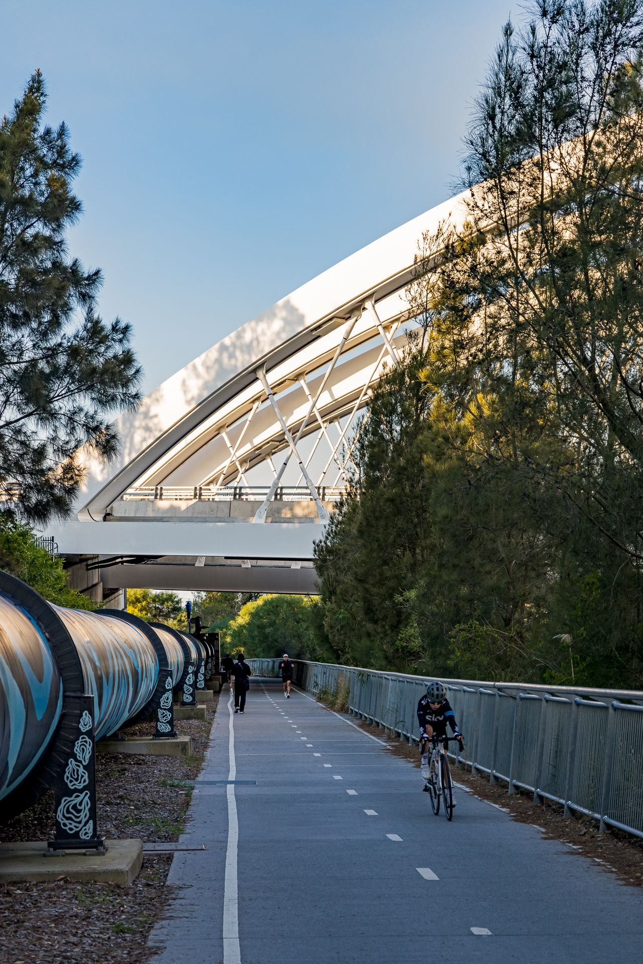 Cyclists and pedestrians on a shared bike path beside a large pipeline, leading toward a white arched bridge.