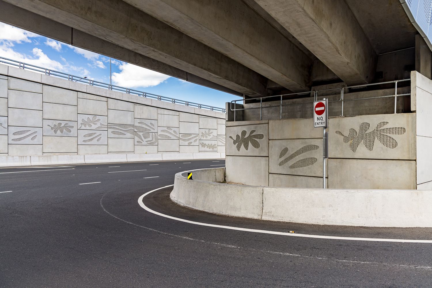 Underside of a concrete overpass with decorative leaf-patterned wall panels beside a curving road with a stop sign on the barrier.