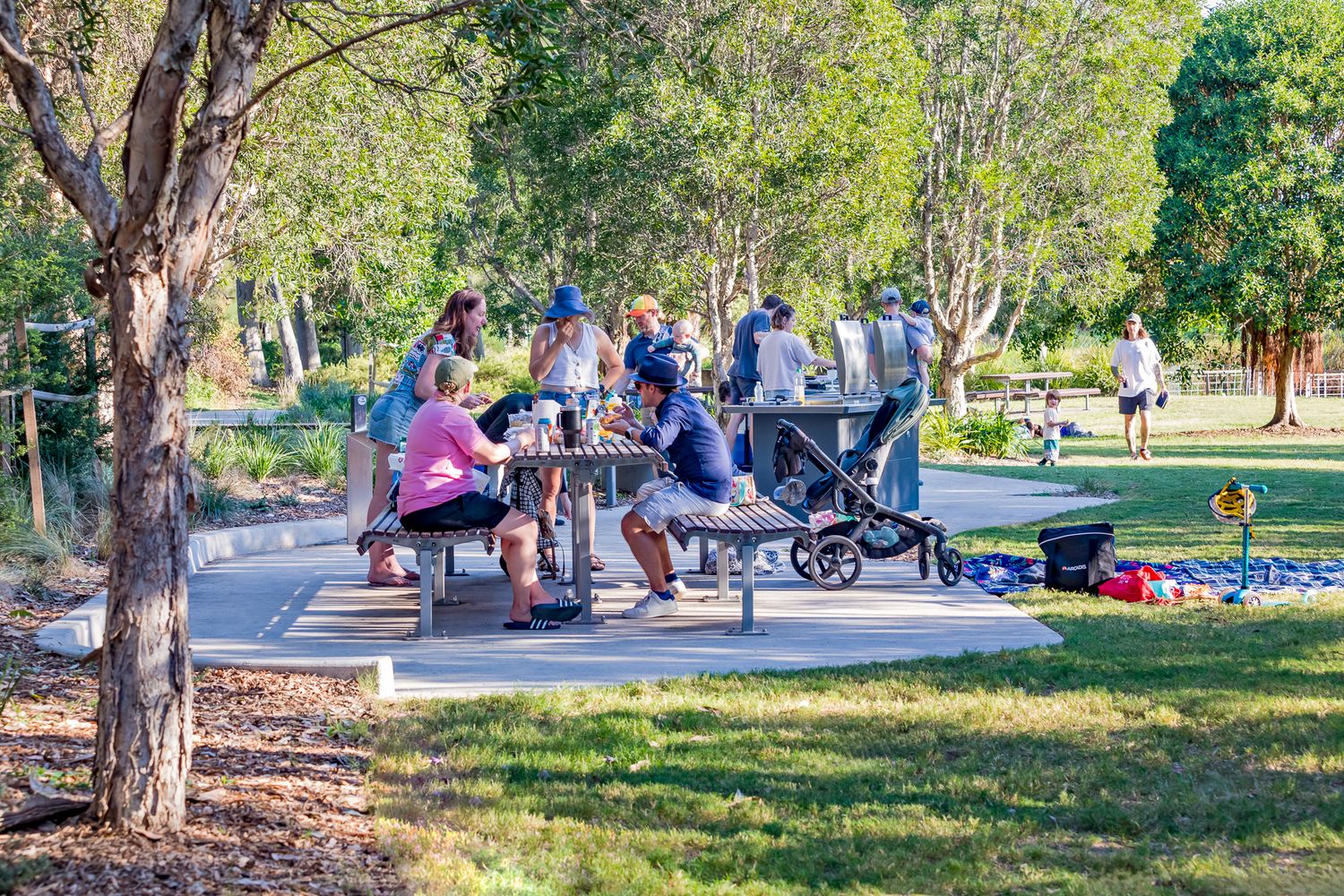 Families and friends enjoy a picnic at tables on a paved area beside a barbecue, with a pram and bags on the lawn in Sydney Park.