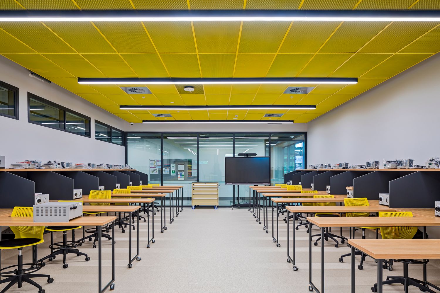 Bright electrical training classroom with rows of workbenches, yellow chairs and a golden ceiling with linear lighting.