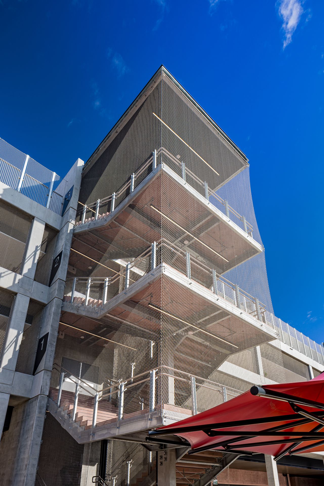 Exterior concrete stairwell with metal mesh cladding and railings rising against a deep blue sky beside a red canopy.