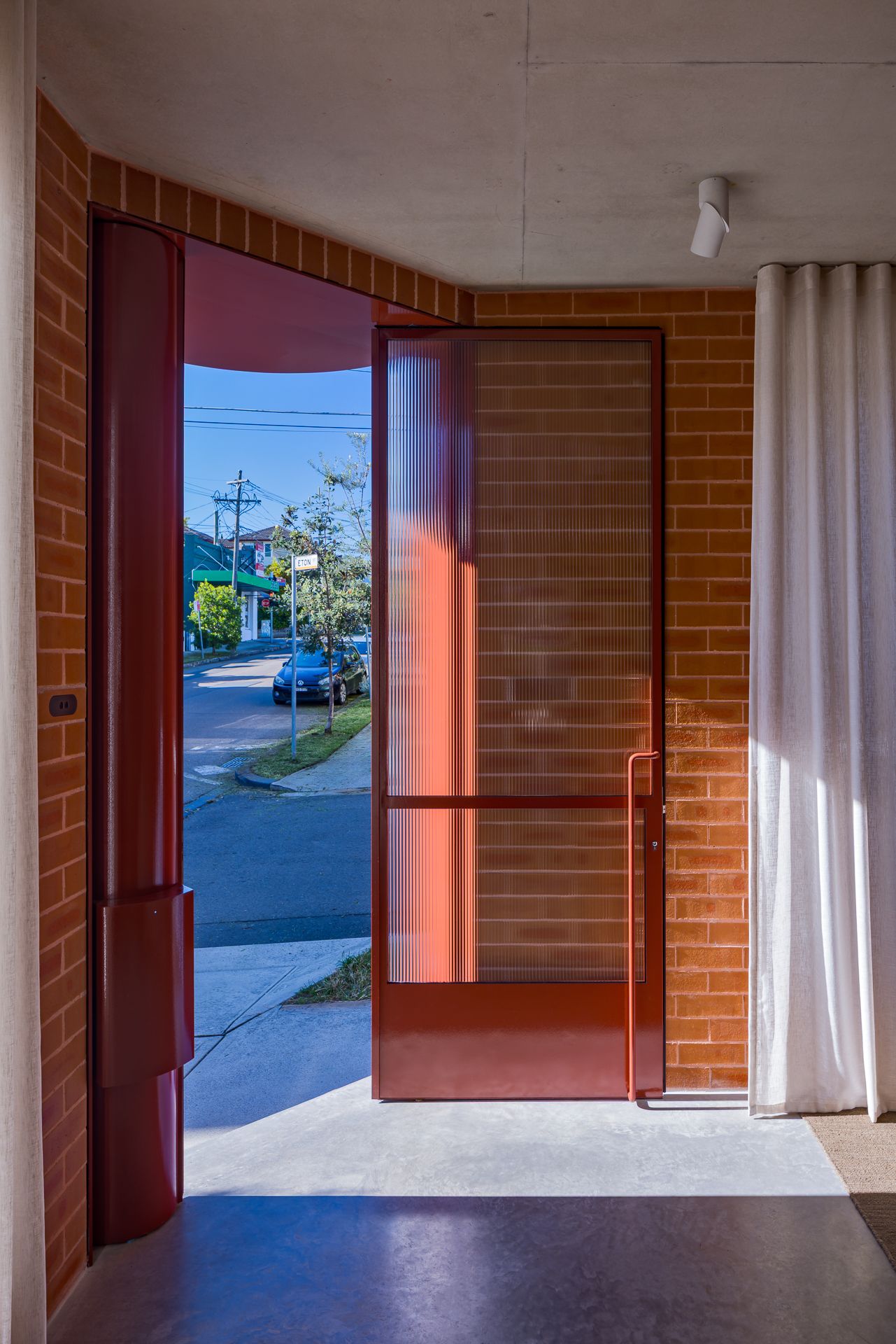 Open red-framed glass entry door in a brick wall, viewed from inside, with a street, trees and white curtain beyond.