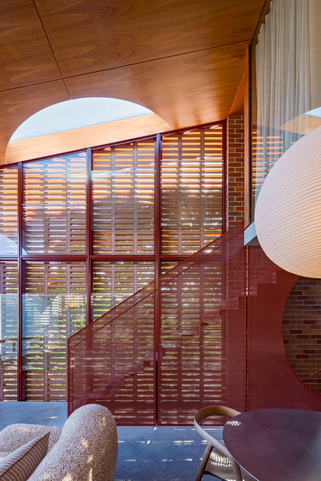 Interior with curved plywood ceiling, red perforated metal staircase, timber louvre screens, brick wall, pendant lamp and bouclé armchair.