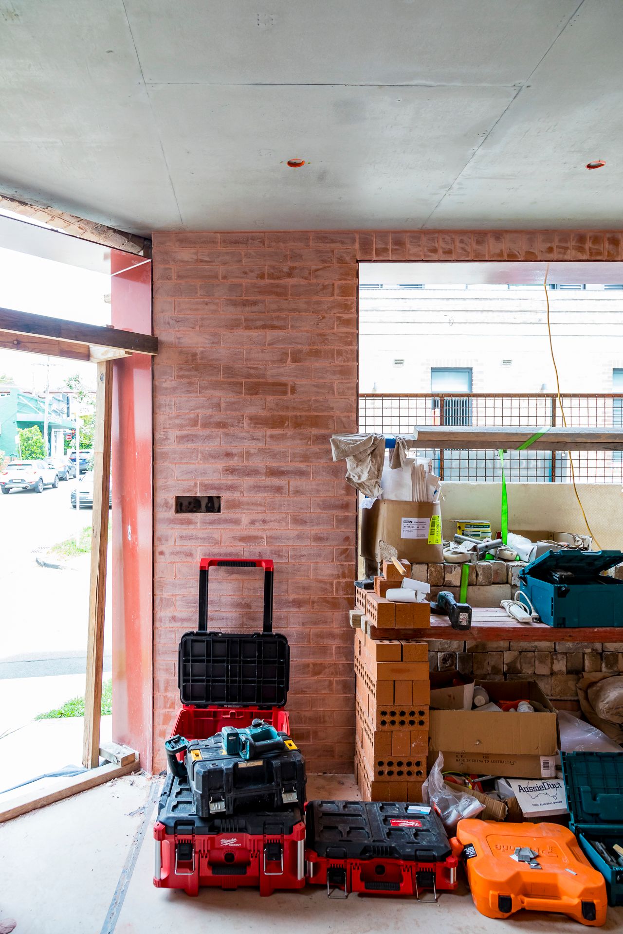 Construction site interior with red toolboxes and supplies in front of a pink brick wall, concrete ceiling and open window.