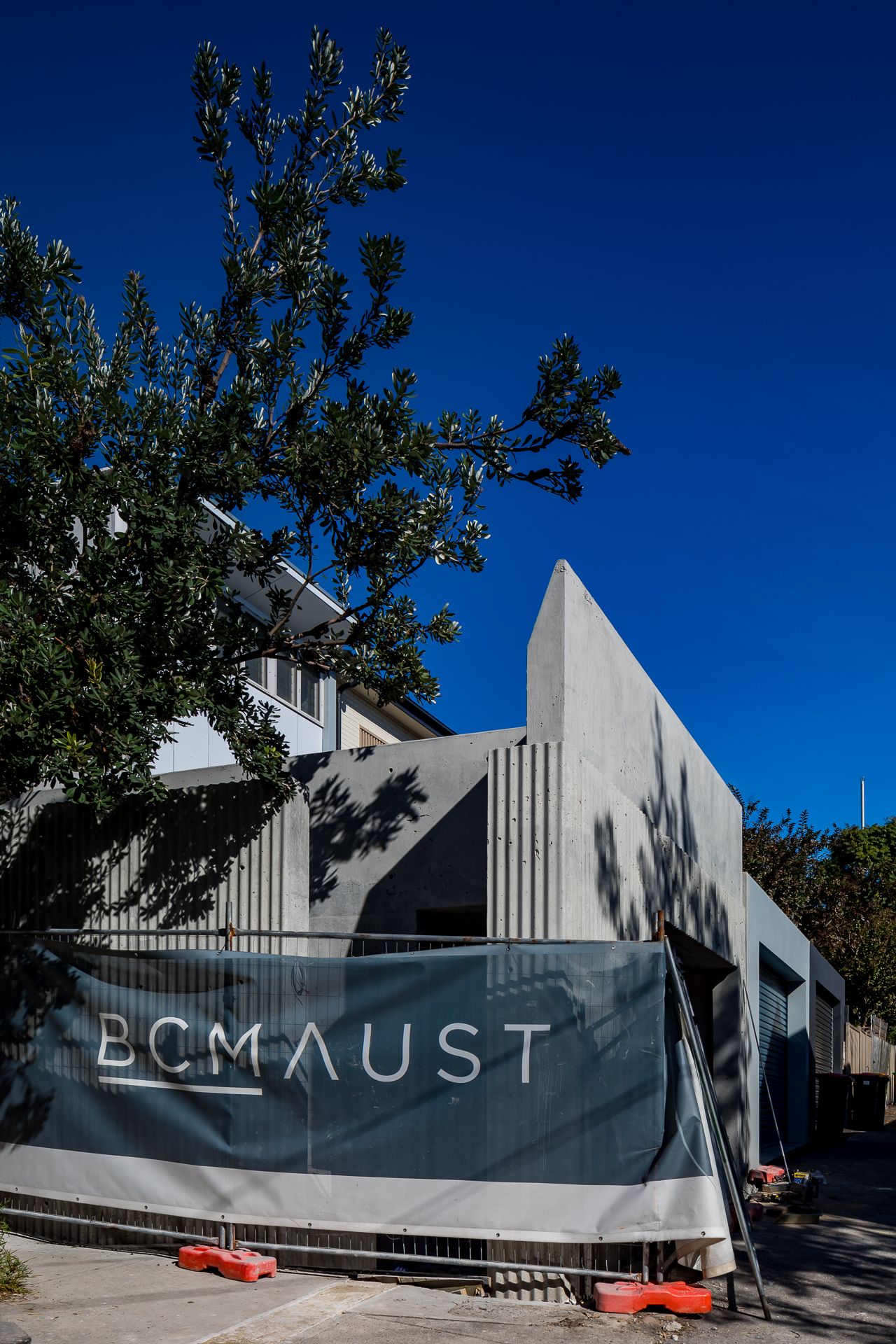 Modern concrete building behind a construction hoarding reading BCM AUST, with a tree and deep blue sky.