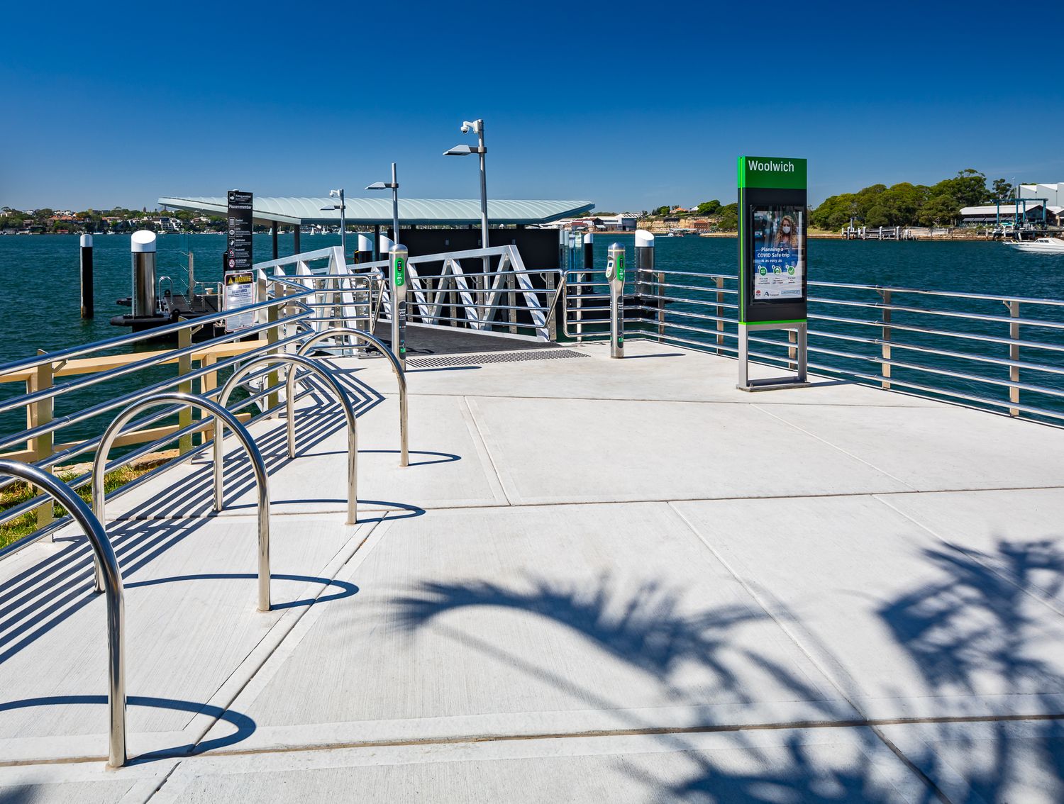 Ferry wharf platform with bike racks, railings and an information sign beside sparkling blue harbour water on a sunny day.
