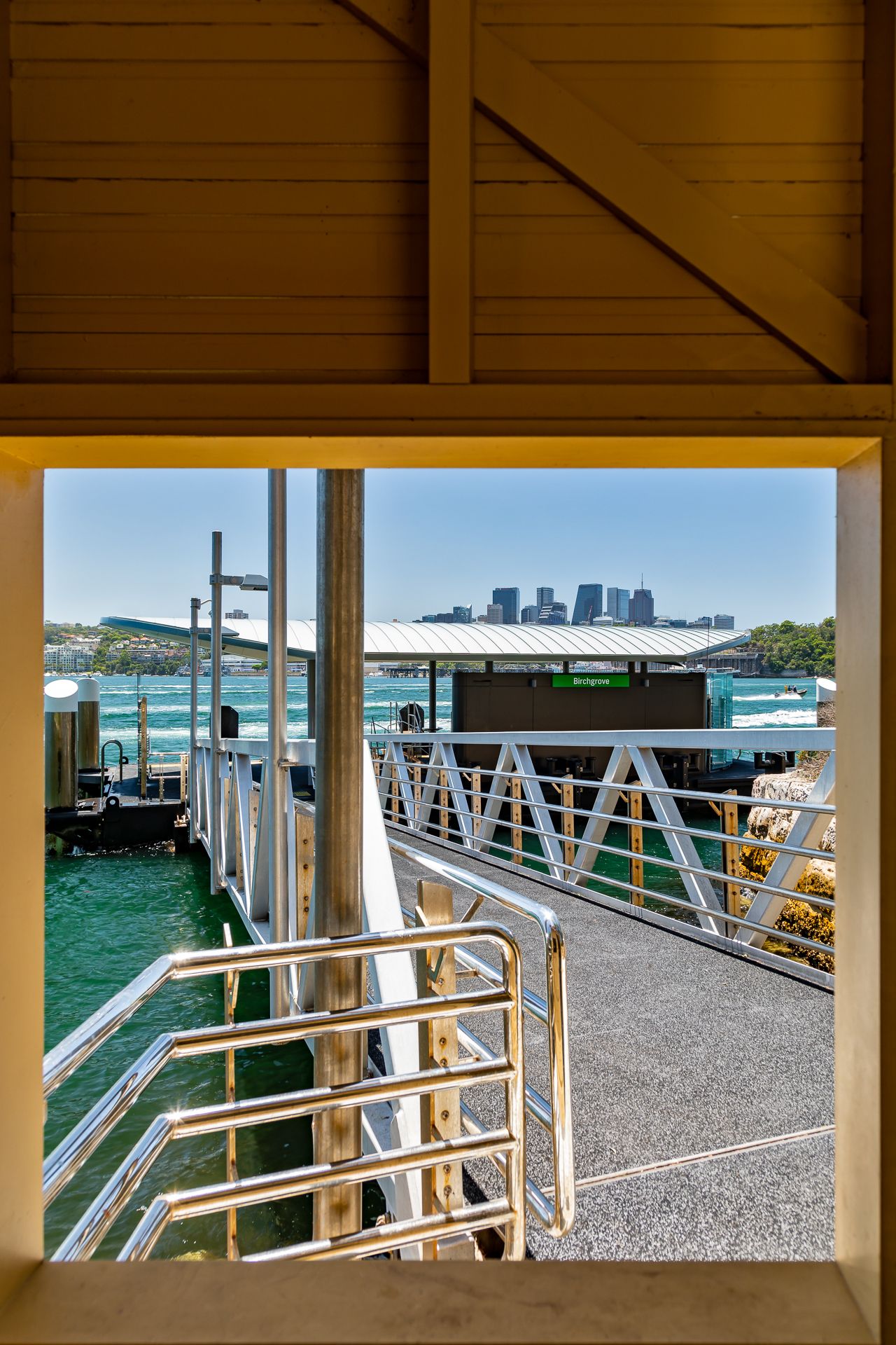View through a timber wharf frame toward a ferry wharf with metal railings, a gangway ramp and a distant city skyline across the water.