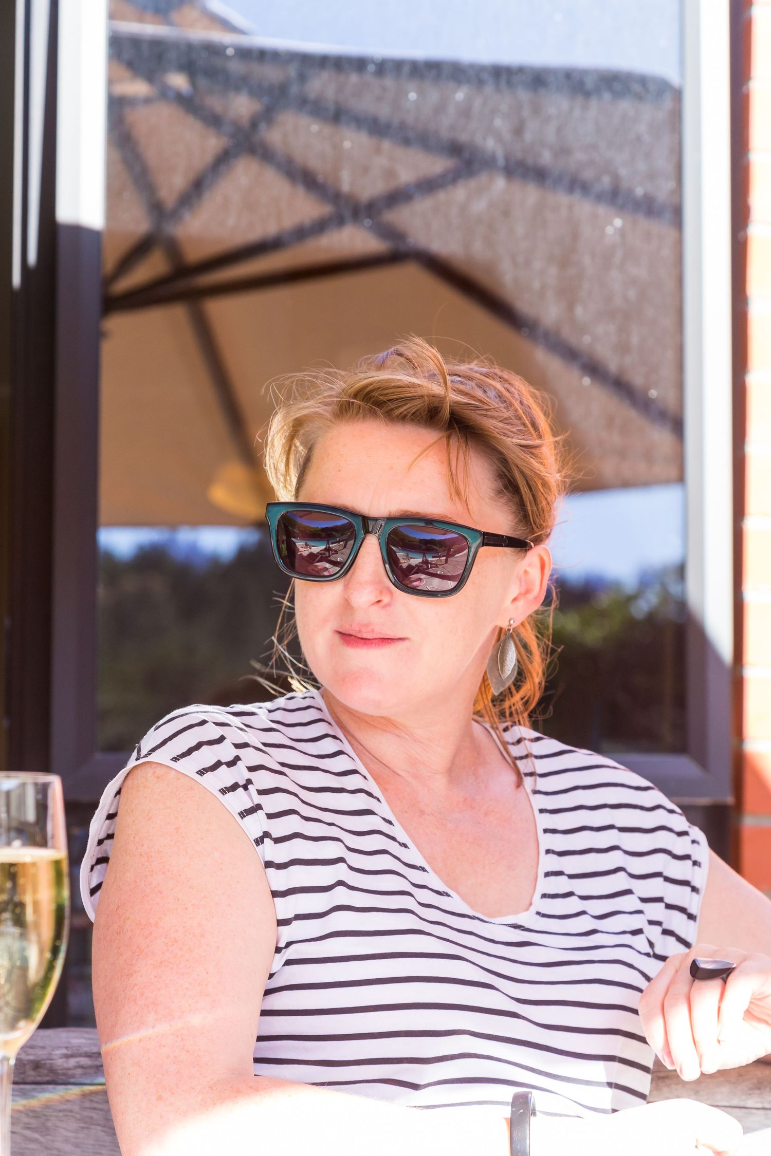 Woman in sunglasses and a striped top sits at an outdoor table beside a glass of sparkling wine on a bright sunny day.