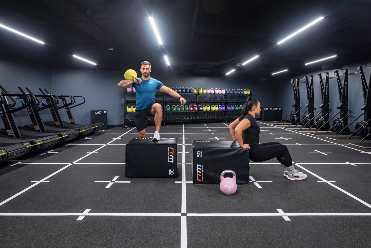 Two people exercising in a modern gym, one holding a medicine ball and the other doing a box exercise beside a kettlebell.