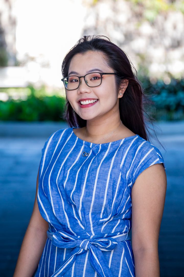 Smiling woman with long dark hair and glasses wears a blue striped dress and poses outdoors with greenery behind her.