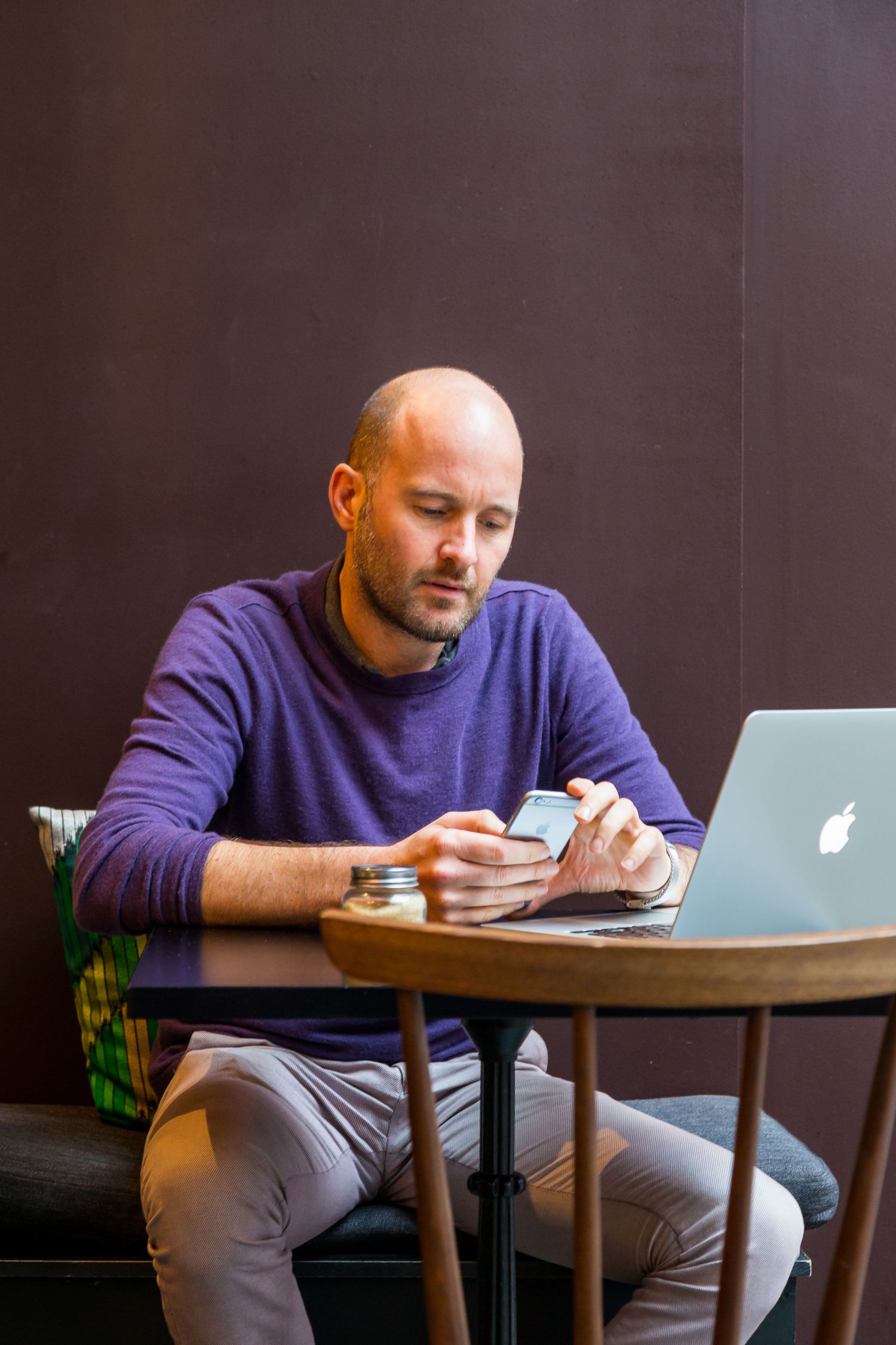 A bald bearded man in a purple sweater checks his phone at a wooden table with an Apple laptop open beside him.