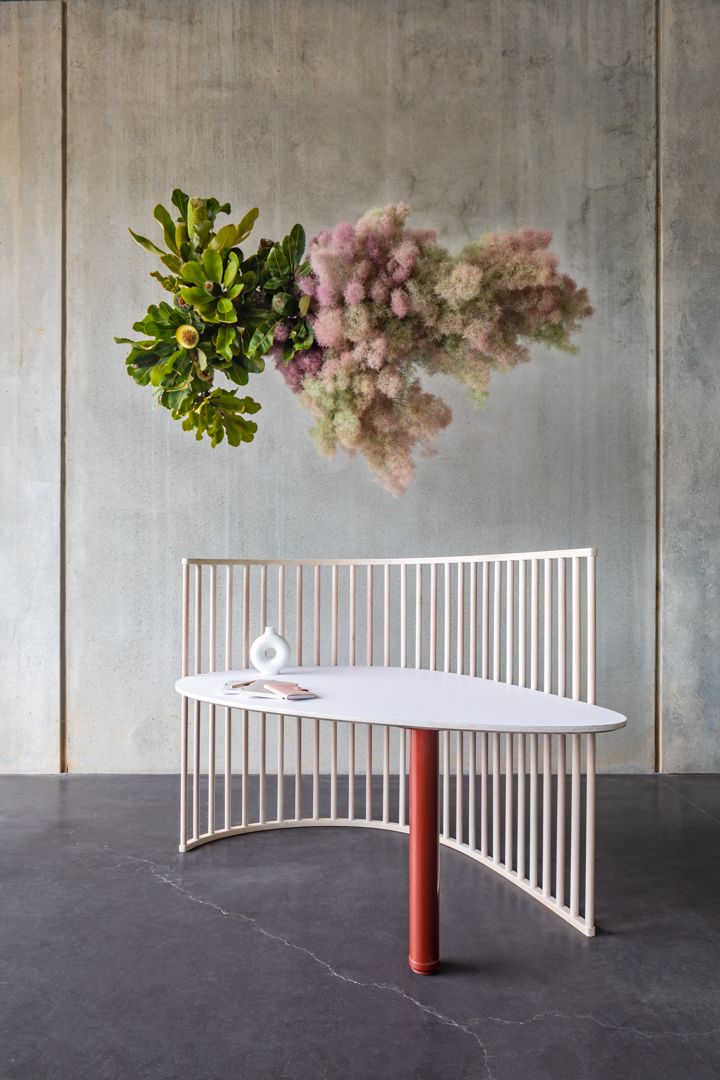 Curved cream slatted bench with white tabletop and red leg beneath a suspended arrangement of green and pink foliage.