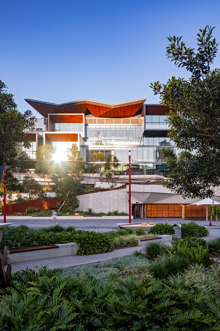 Modern convention centre with a sweeping timber roof and glass facade viewed through lush green landscaping at sunset.