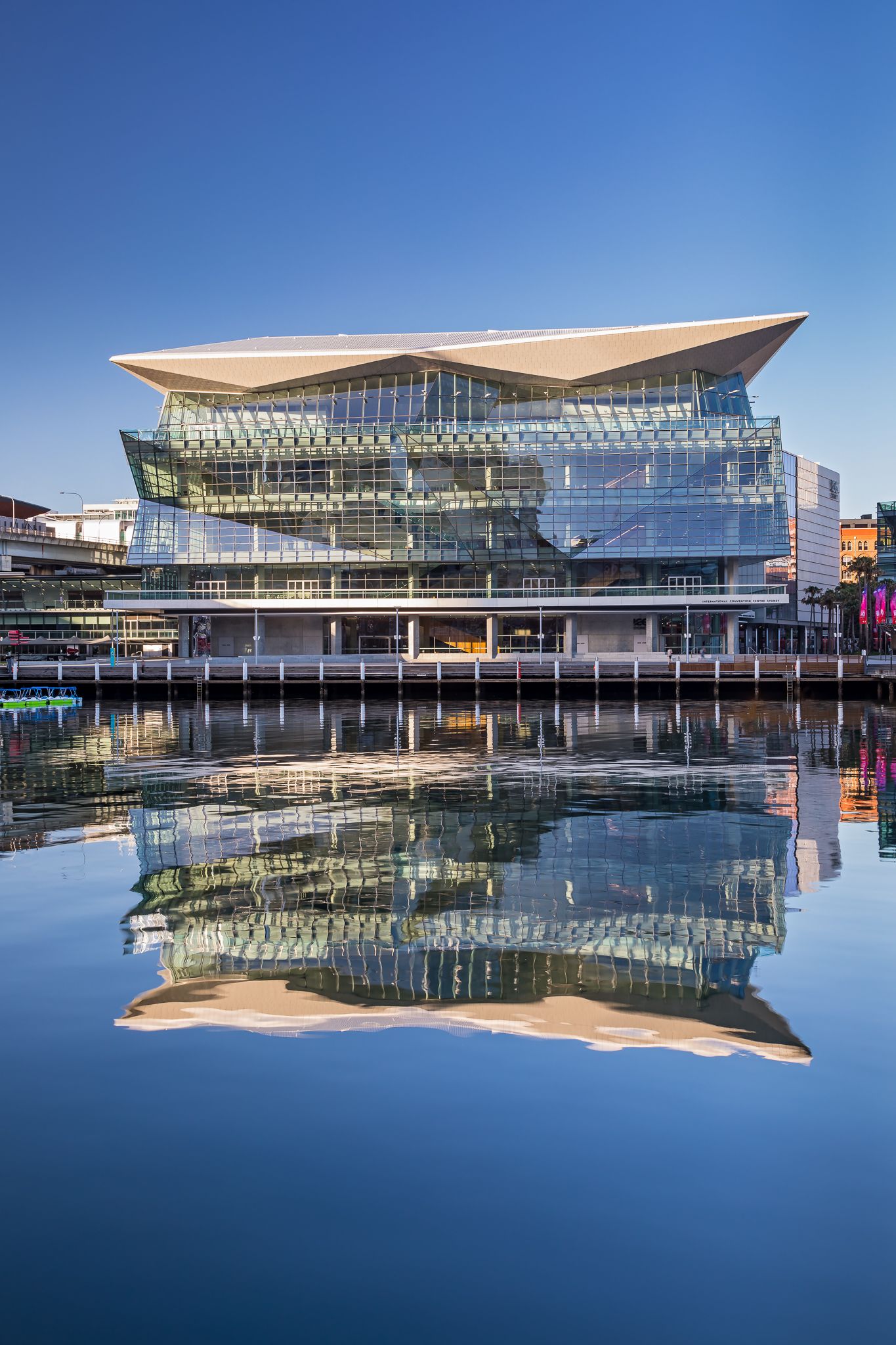 Modern convention centre with a sweeping angular roof and glass facade reflected in calm blue harbour water under a clear sky.