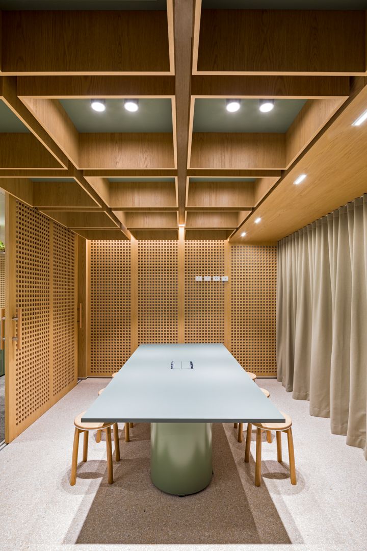 Modern meeting room with timber lattice ceiling, perforated wood wall panels, curtain and a pale green table with wooden stools.