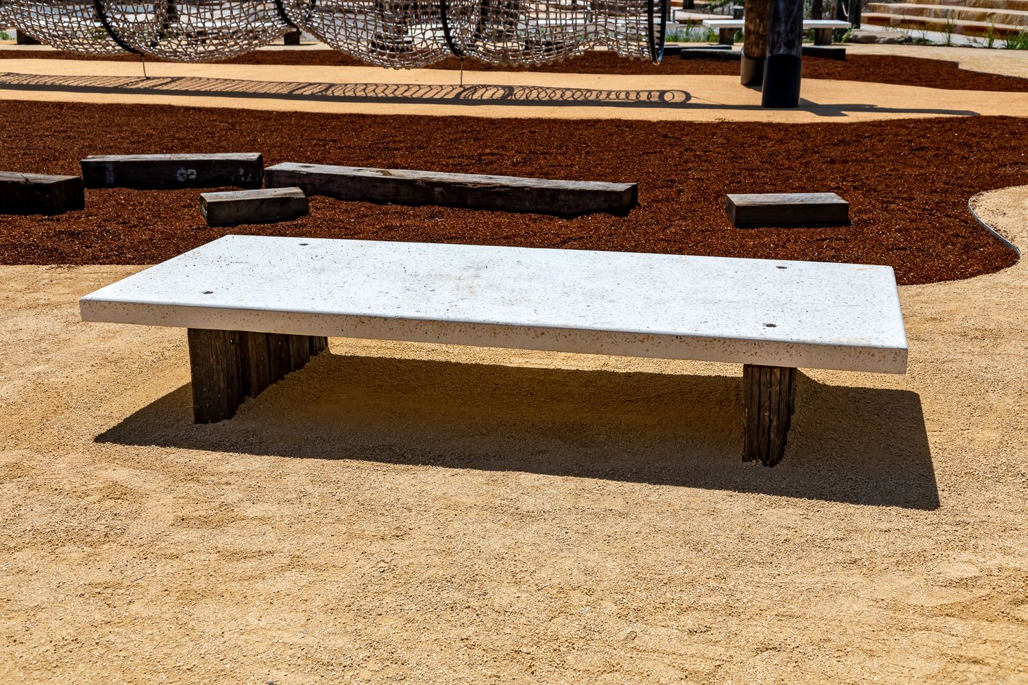 Pale concrete bench on timber legs casting a shadow beside red mulch garden beds and sandy paving in a public park.