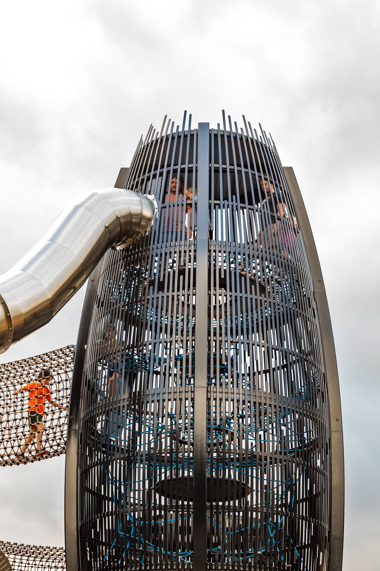 Children climb a tall steel mesh playground tower with rope nets, a silver slide exit, and a net bridge on the left.