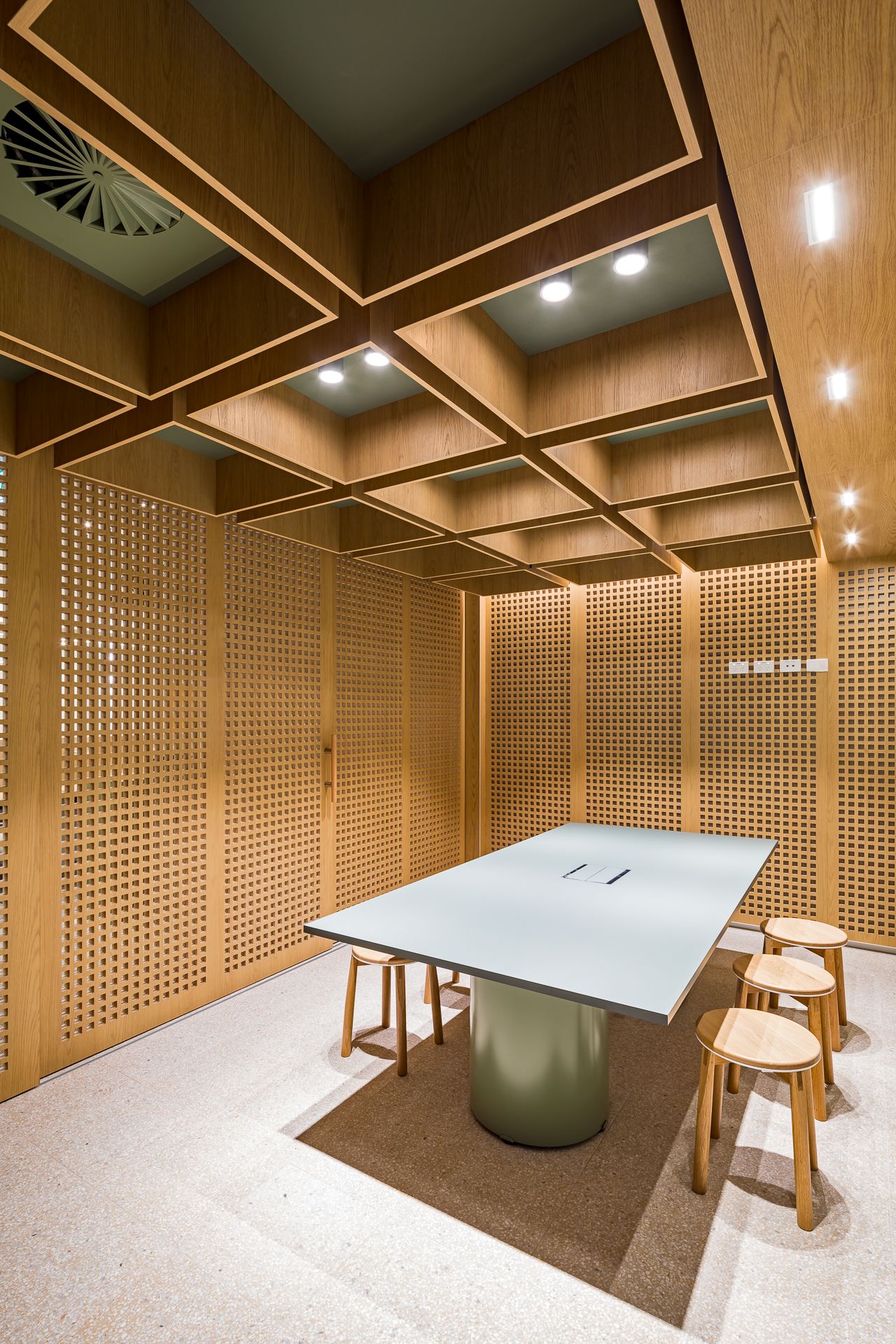 Timber-clad meeting room with perforated wood walls, a coffered ceiling, a gray-green table and round wooden stools.
