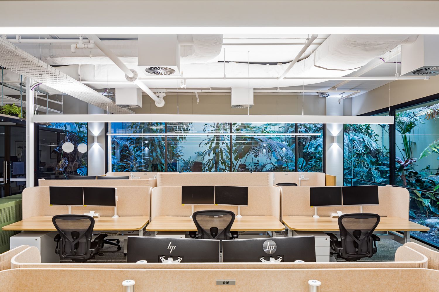 Office workstations with dual monitors and ergonomic chairs facing glass windows that overlook a lush indoor garden courtyard.