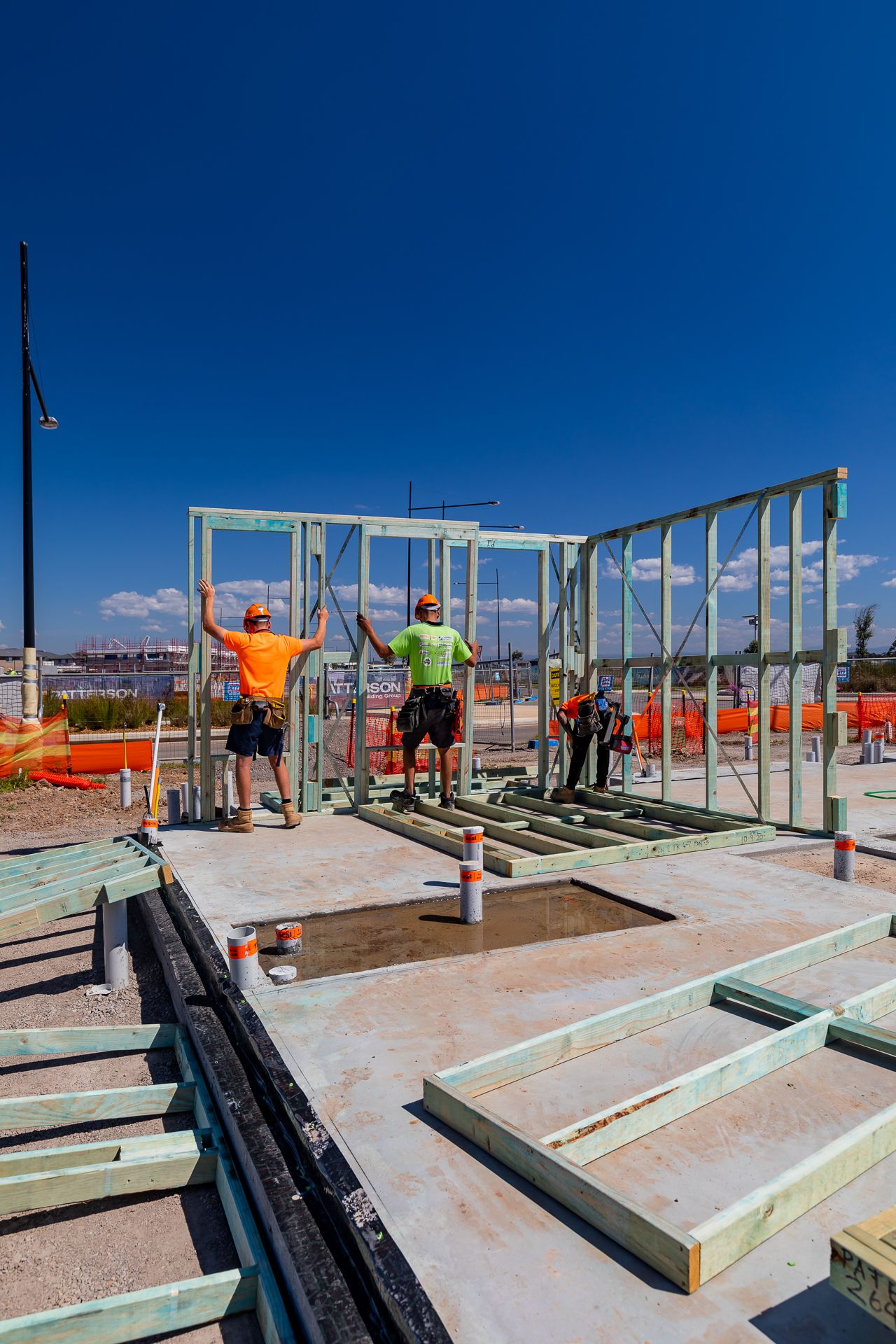 Builders erect a timber wall frame on a concrete slab at a residential construction site on a bright sunny day.
