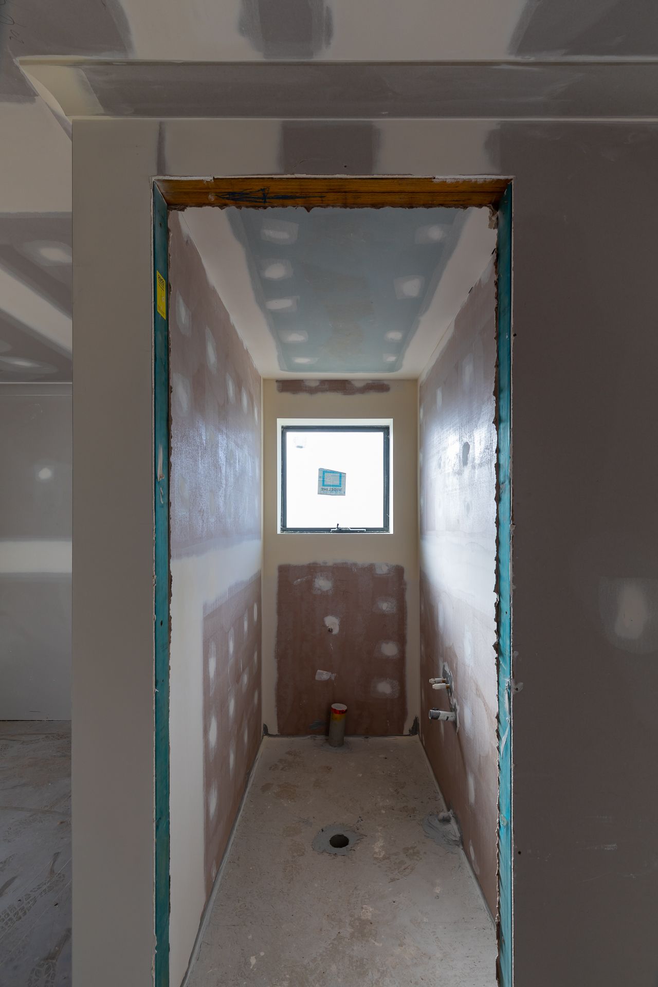 Narrow unfinished bathroom with plasterboard walls, exposed concrete floor, and a small window at the end of the room.