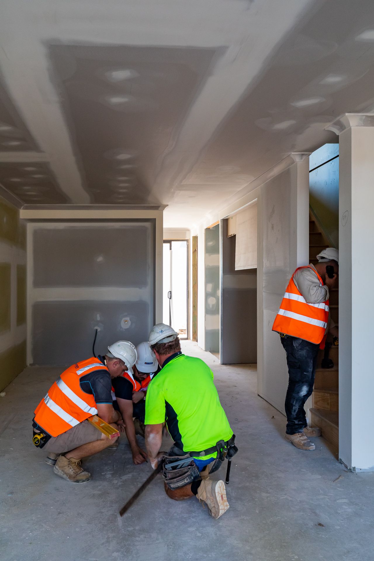 Construction workers in hi-vis vests and hard hats inspect plasterboard walls and ceilings inside a new home under construction.