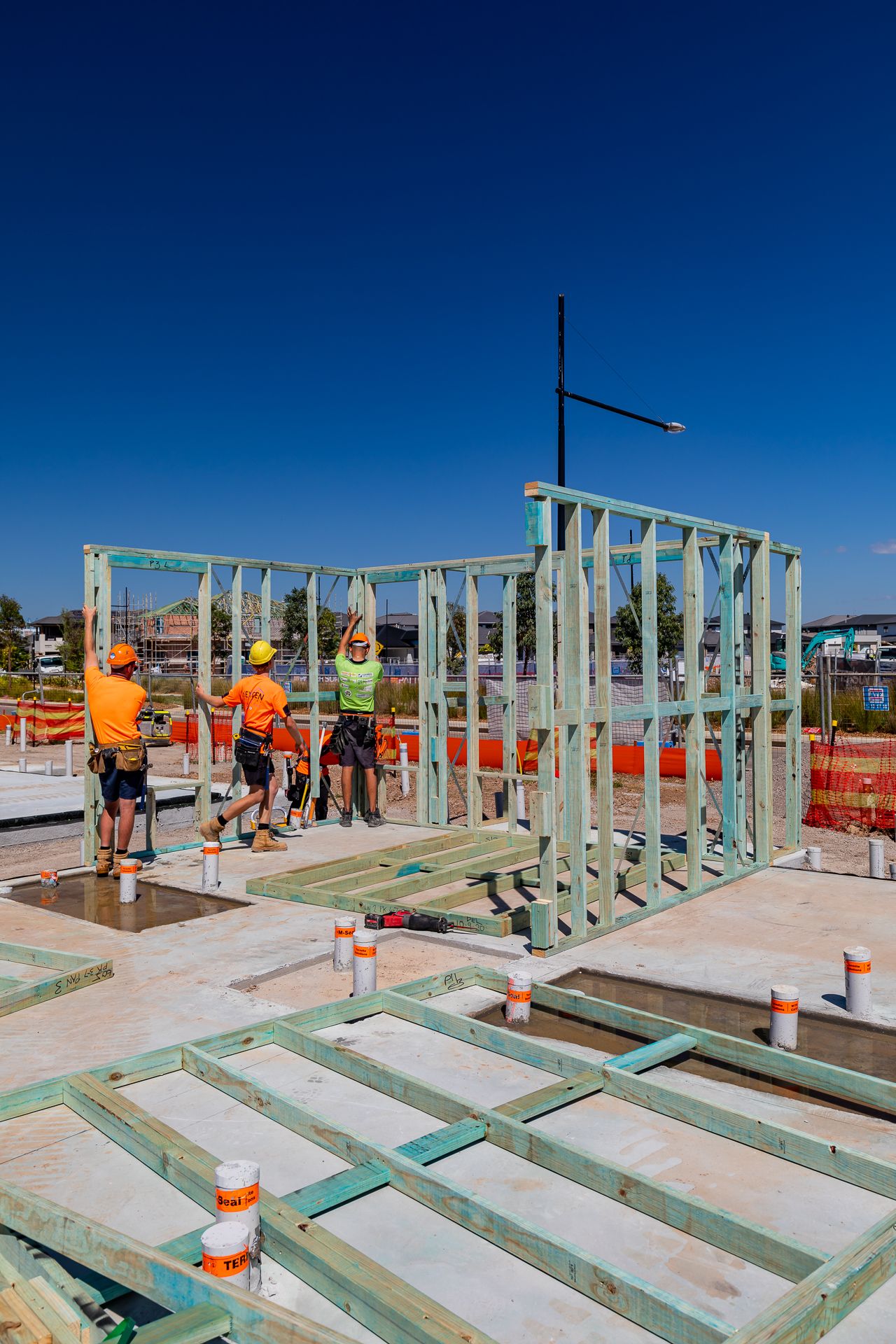 Three workers raise a steel wall frame on a concrete slab with plumbing pipes and floor framing in the foreground.