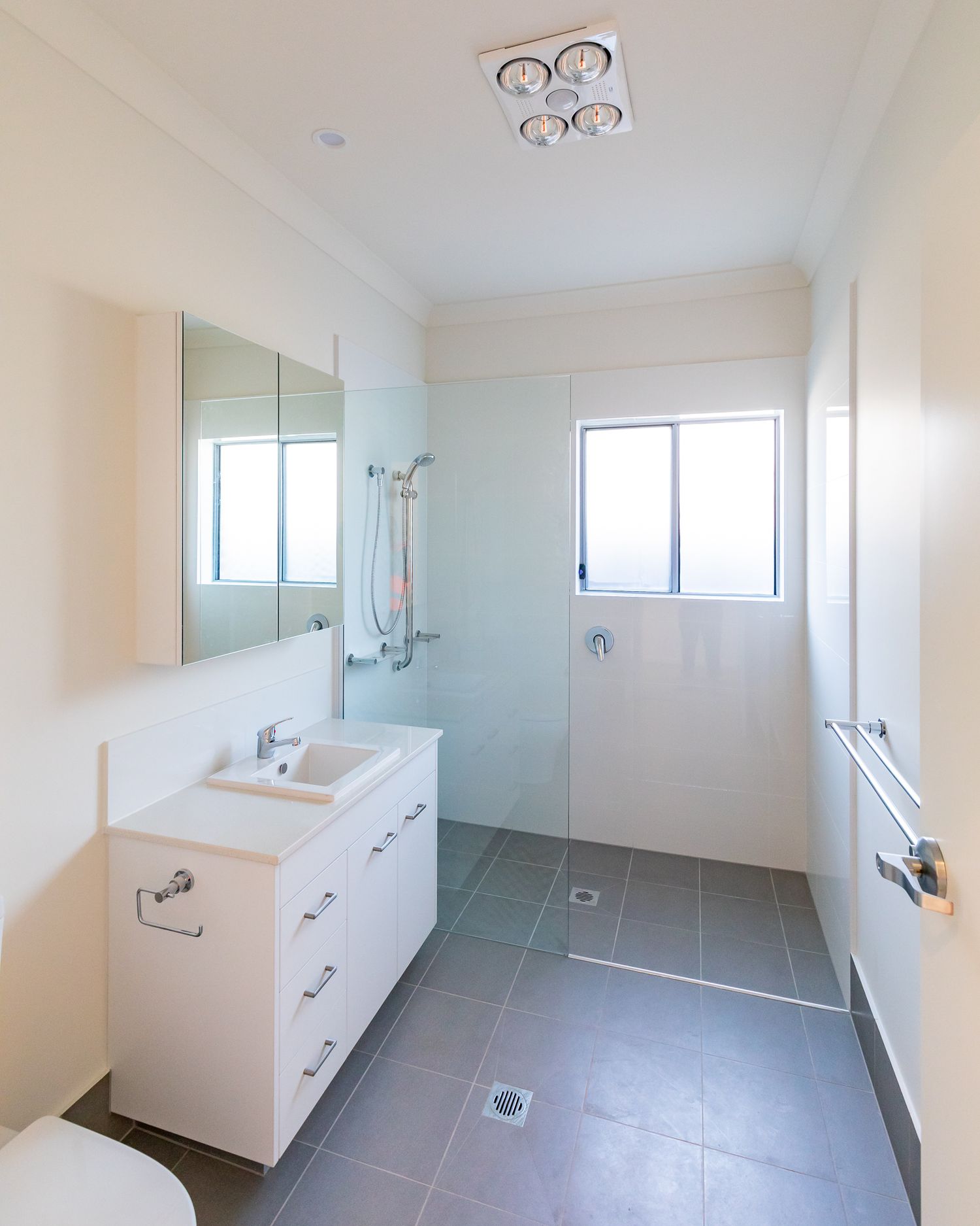 Bright modern bathroom with white vanity, mirrored cabinet, glass shower screen, heat lamp and grey floor tiles.