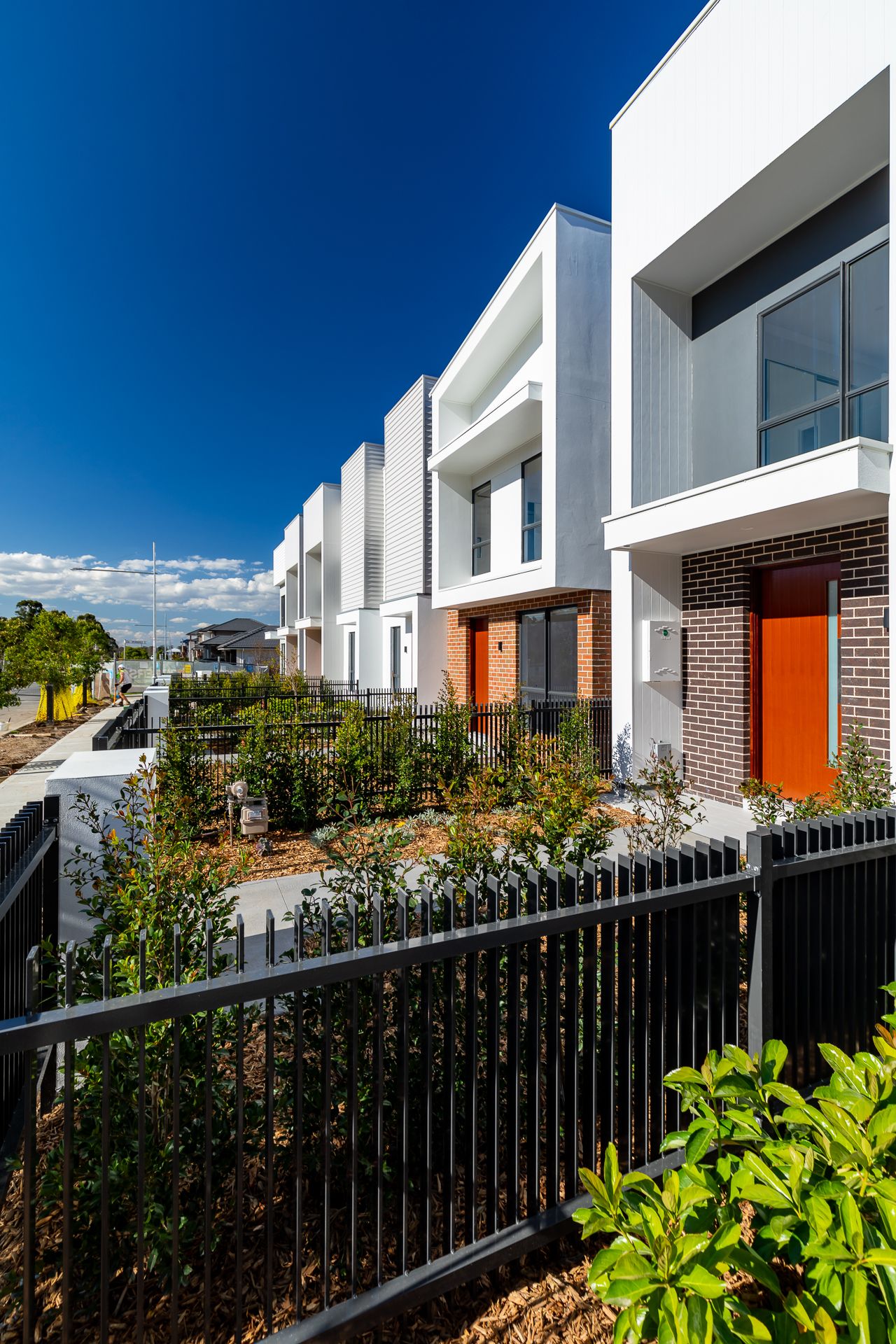 Modern townhouse row with white facades, orange front doors, brick details and new landscaping behind a black fence.