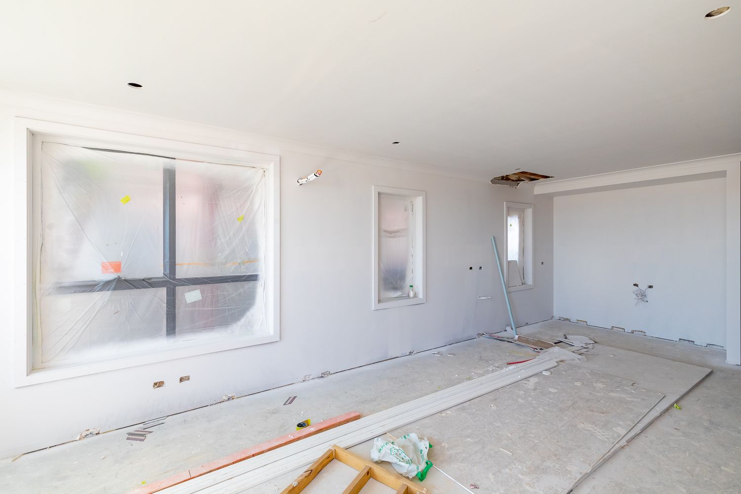 Unfinished room in a new townhouse with plastered walls, large windows covered in protective film and a dusty concrete floor.