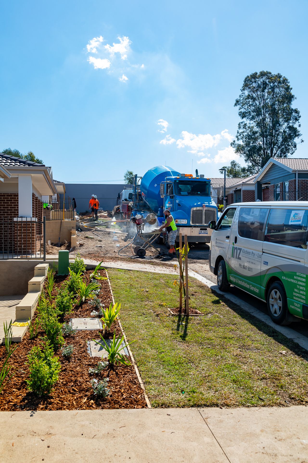 Concrete mixer truck and workers pour concrete at a residential construction site beside a freshly landscaped garden and a parked work van.