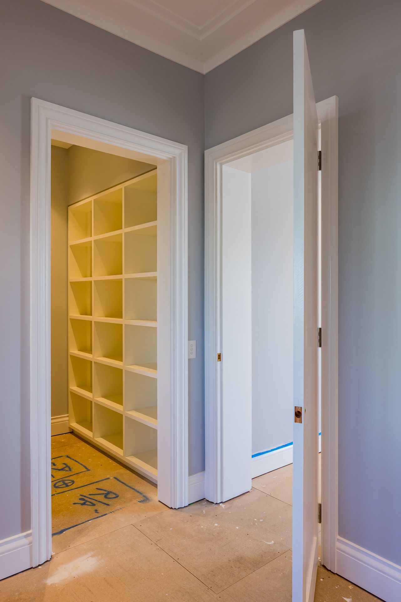 Hallway with an open white door revealing a walk-in storage room fitted with empty white shelving and a stone-tiled floor.