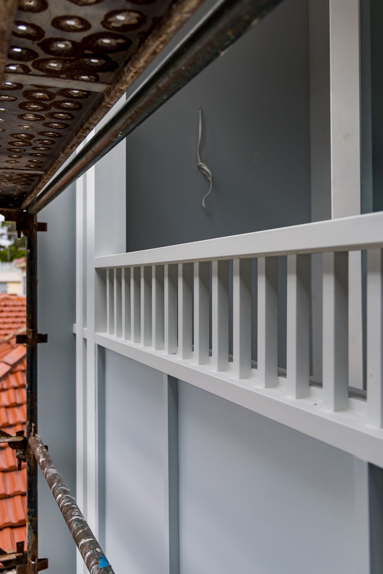 Exterior view of a blue-grey wall with a white timber balustrade and scaffolding beside terracotta roof tiles.