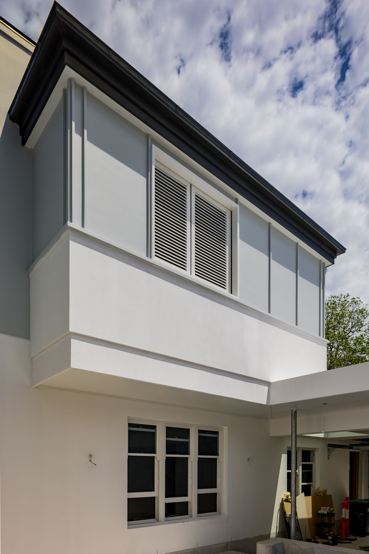 Exterior of a renovated white home with a dark roof trim, louvered shuttered windows on an upper level and a black-framed window below.