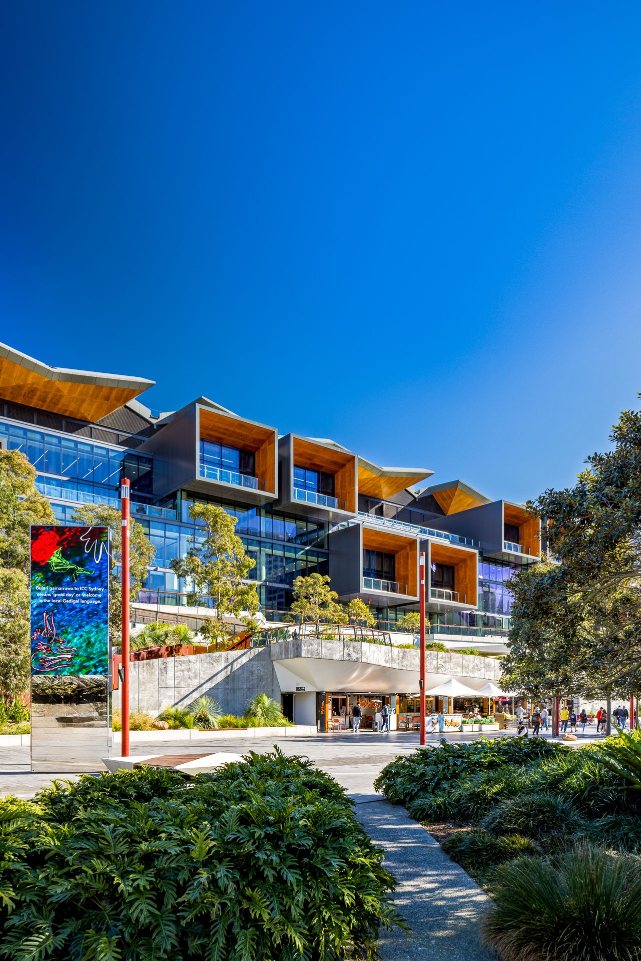 Multi-storey exhibition centre at ICC Sydney with timber-lined balconies, native plantings and a pedestrian path beneath a bright blue sky.