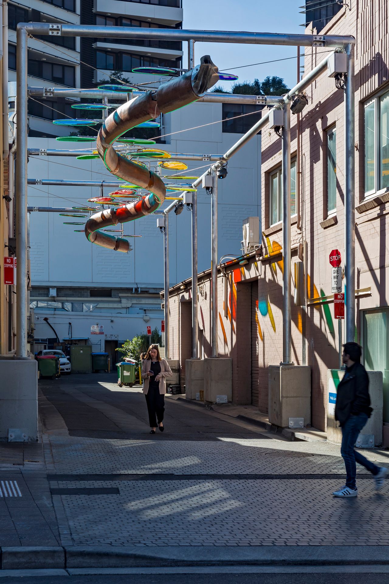 Woman walks down a sunlit alley beneath a hanging spiral art installation of multicolored discs and a brick building.