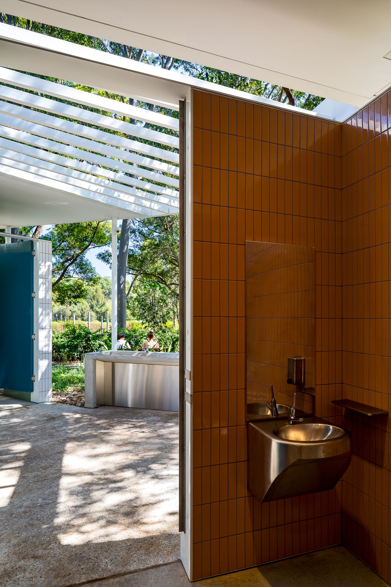 Interior of a park restroom with warm orange tiled walls, a stainless steel wall-mounted basin and views of greenery.