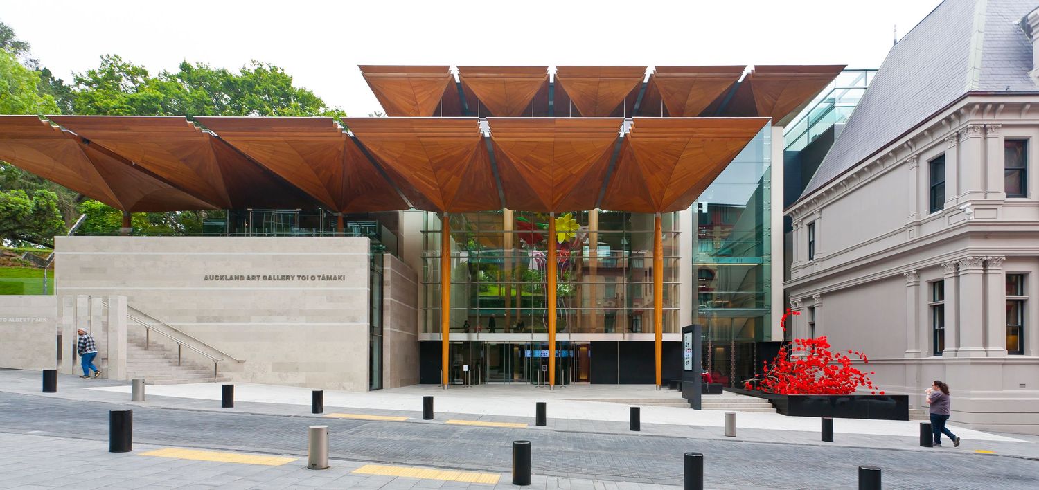 Modern art gallery entrance with a timber lattice canopy, glass facade, and stone wall beside a heritage building and red sculpture.