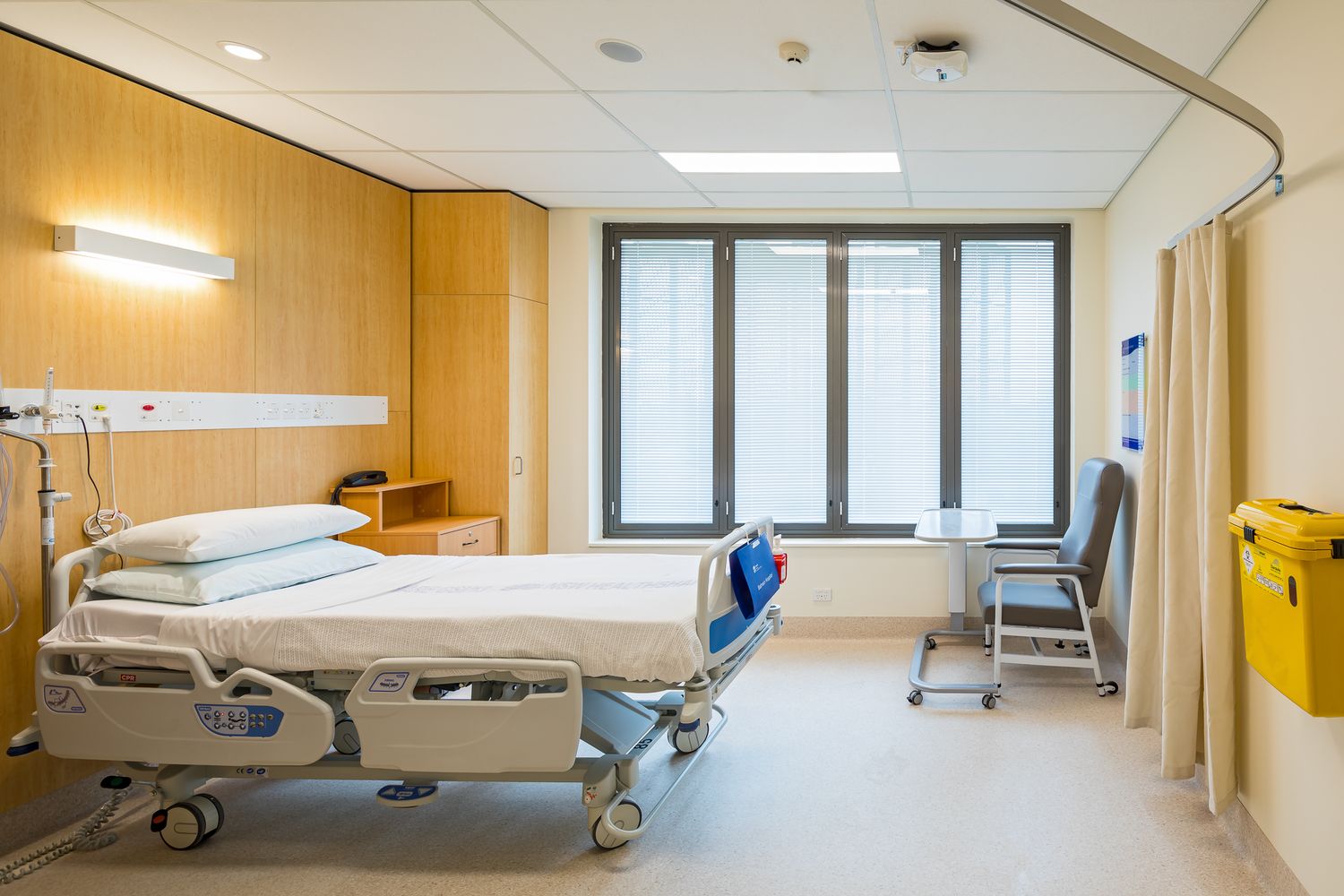 Bright hospital patient room with an adjustable bed, wood paneling, a large window with blinds and a reclining visitor chair.