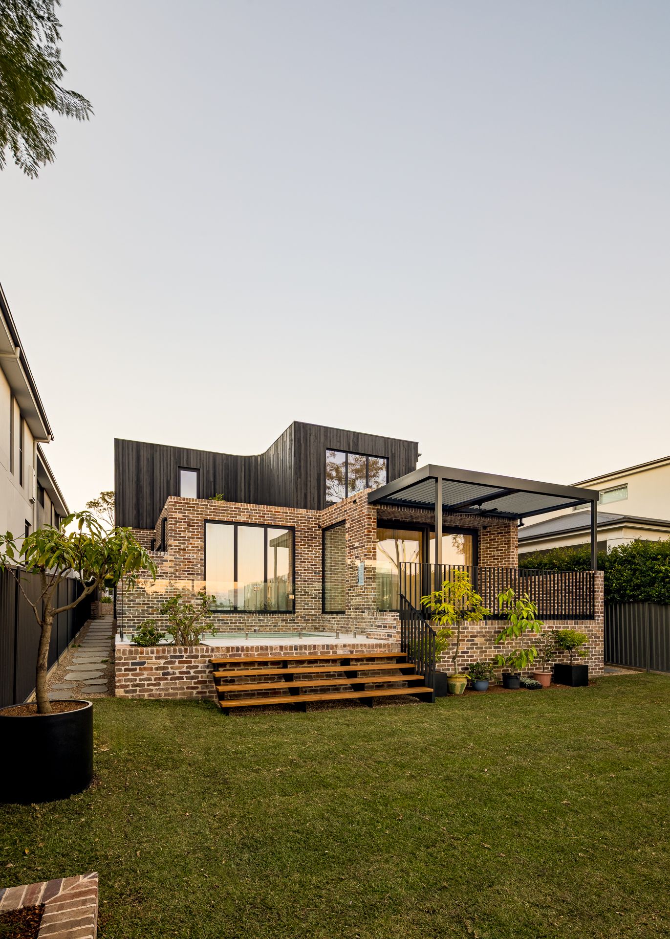 Modern two-storey home with brick base and charred black timber upper level, timber deck steps and a pergola over a landscaped lawn.