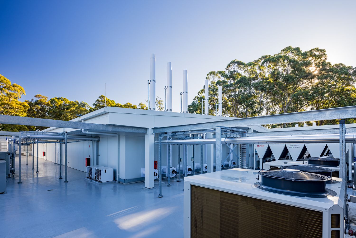 Rooftop plant area with white exhaust stacks, pipework and air conditioning units set against a blue sky and surrounding eucalyptus trees.