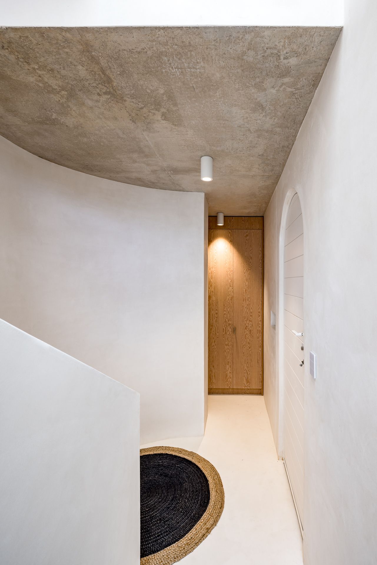 Minimalist hallway with a raw concrete ceiling, curved plaster walls, an arched door, oak panel and a round black woven rug.