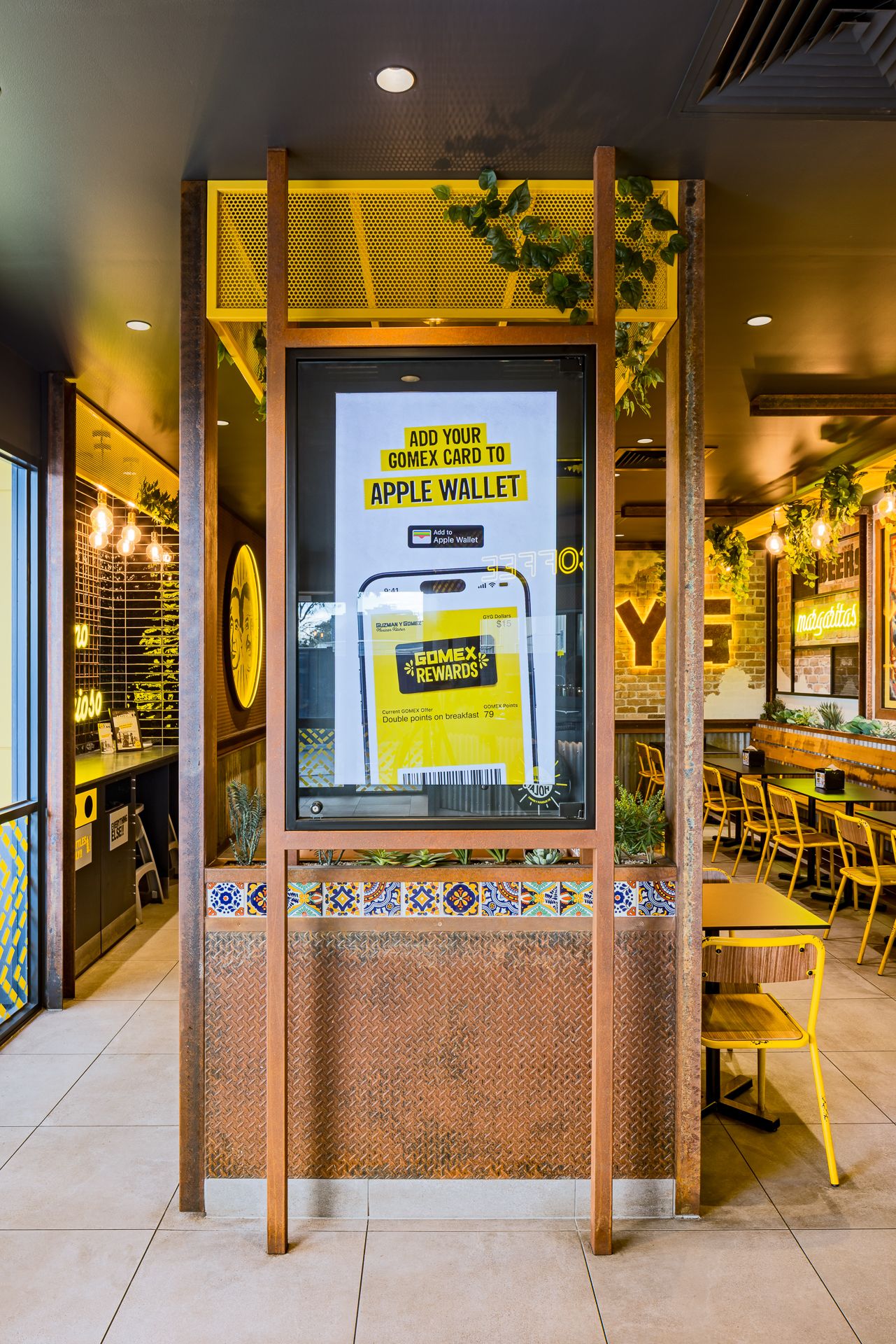 Restaurant interior with a digital screen promoting adding the Gomex rewards card to Apple Wallet beside tiled planters and yellow chairs.