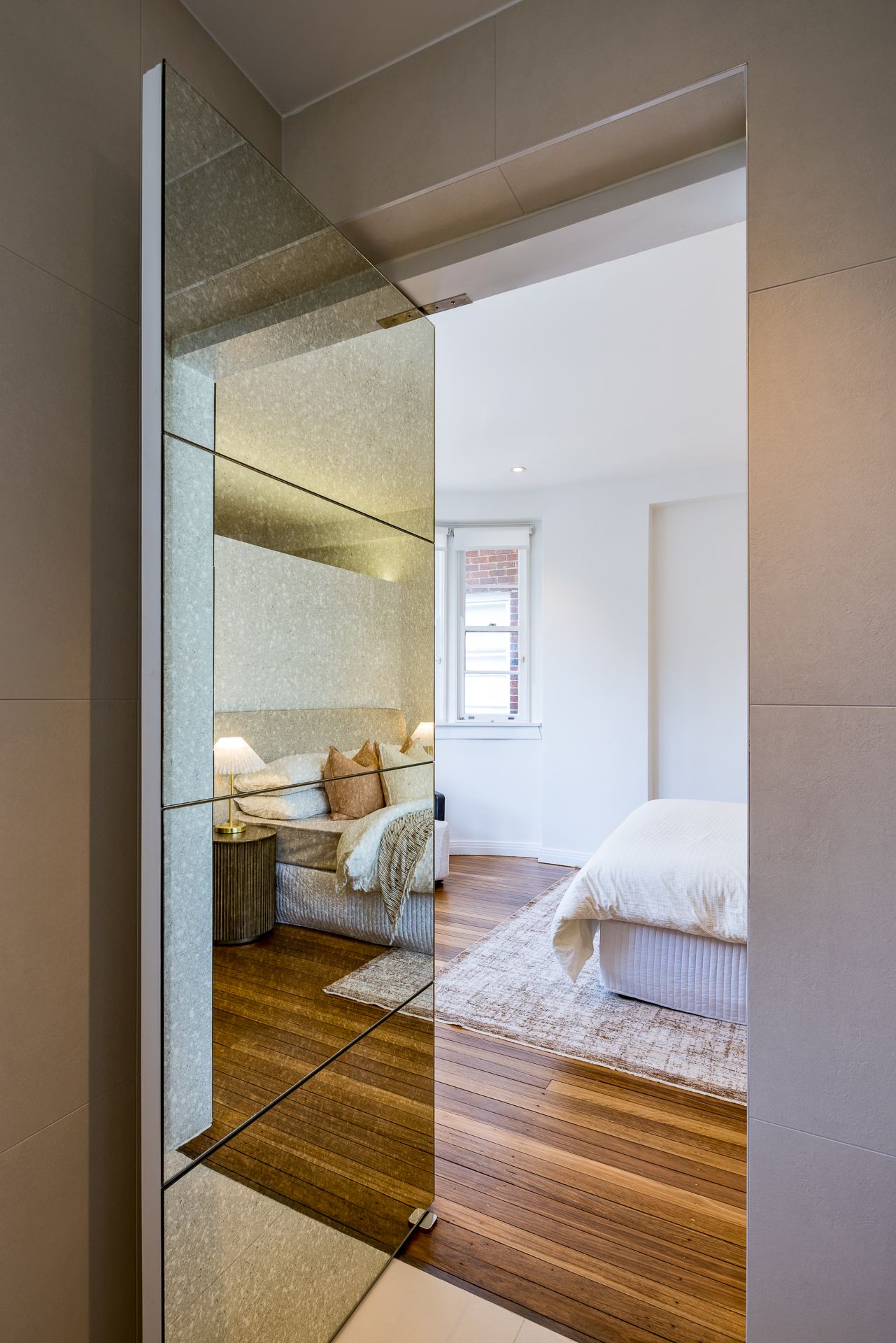 Mirrored pivot door opening to a bright bedroom with timber floors, a neutral rug and a bed with white linen.